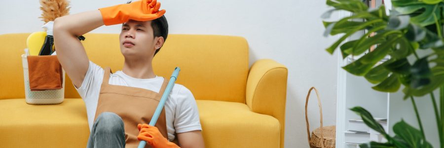 Young housekeeper man sitting near sofa with feel tired, he closed eyes for rest in living room, copy space